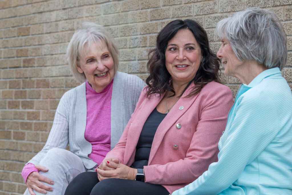 Rep. Witwer sitting between two older women and talking, wearing a pink blazer.